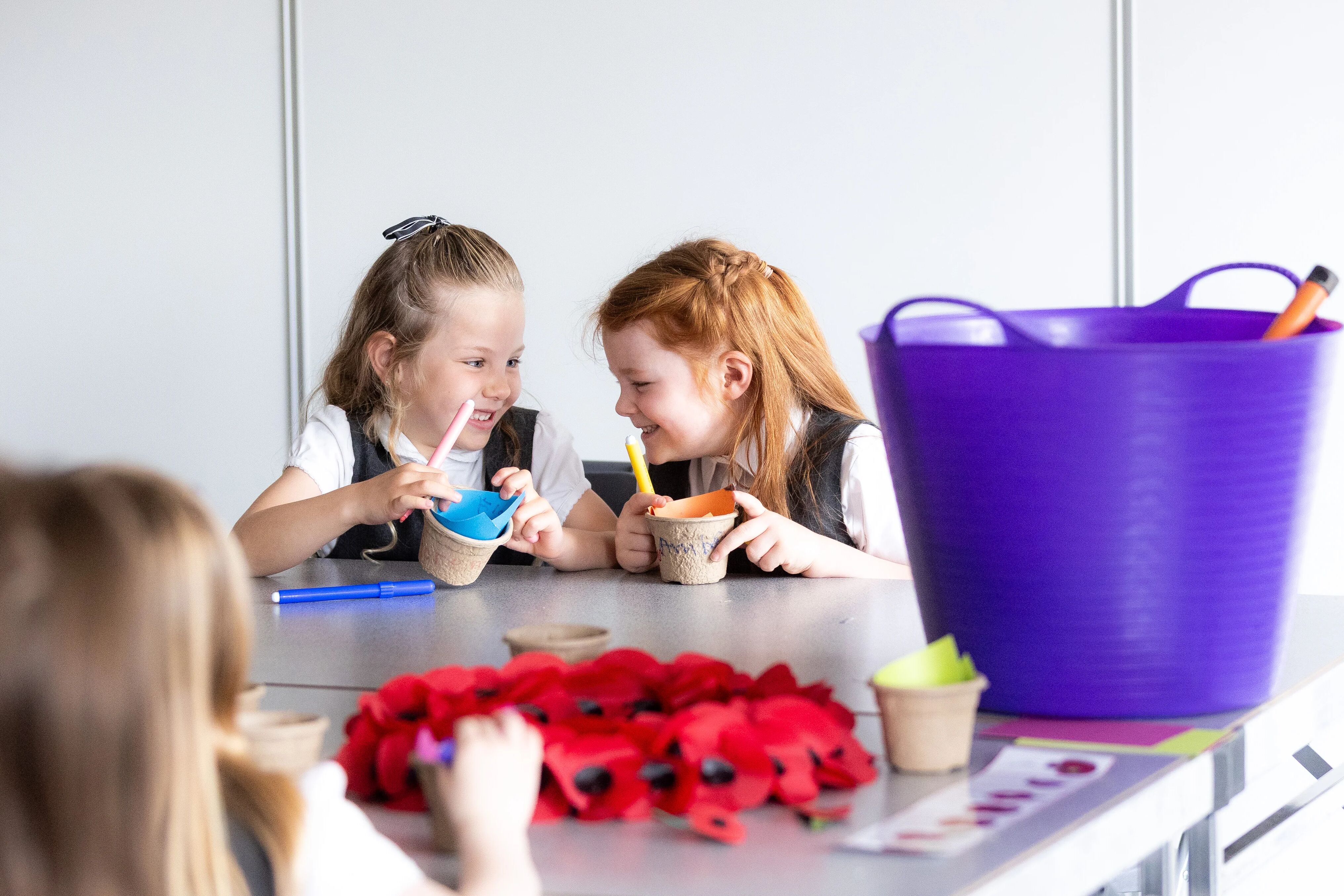 Two girls in key stage one, laughing together while they decorate paper plant pots to plant poppies in as part of an Activity Day at the Arboretum.